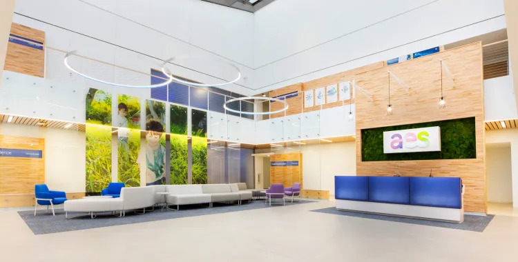 Modern office lobby with high ceilings, circular hanging lights, blue and white seating, wooden wall accents, and a reception desk with a colorful 'aes' logo. Large wall panels display photos of children in nature, creating a welcoming and vibrant atmosphere.