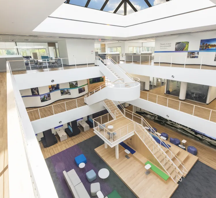 Bright, modern office atrium with open seating areas, wooden floors, white railings, a central staircase, and a large skylight providing natural light. Colorful furniture and wall art create a welcoming, collaborative workspace.