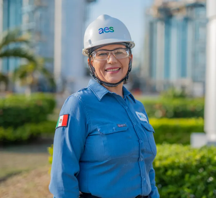 Smiling female engineer wearing safety helmet and glasses, dressed in a blue shirt with a Mexican flag patch, standing outdoors at an industrial site with greenery and buildings in the background.